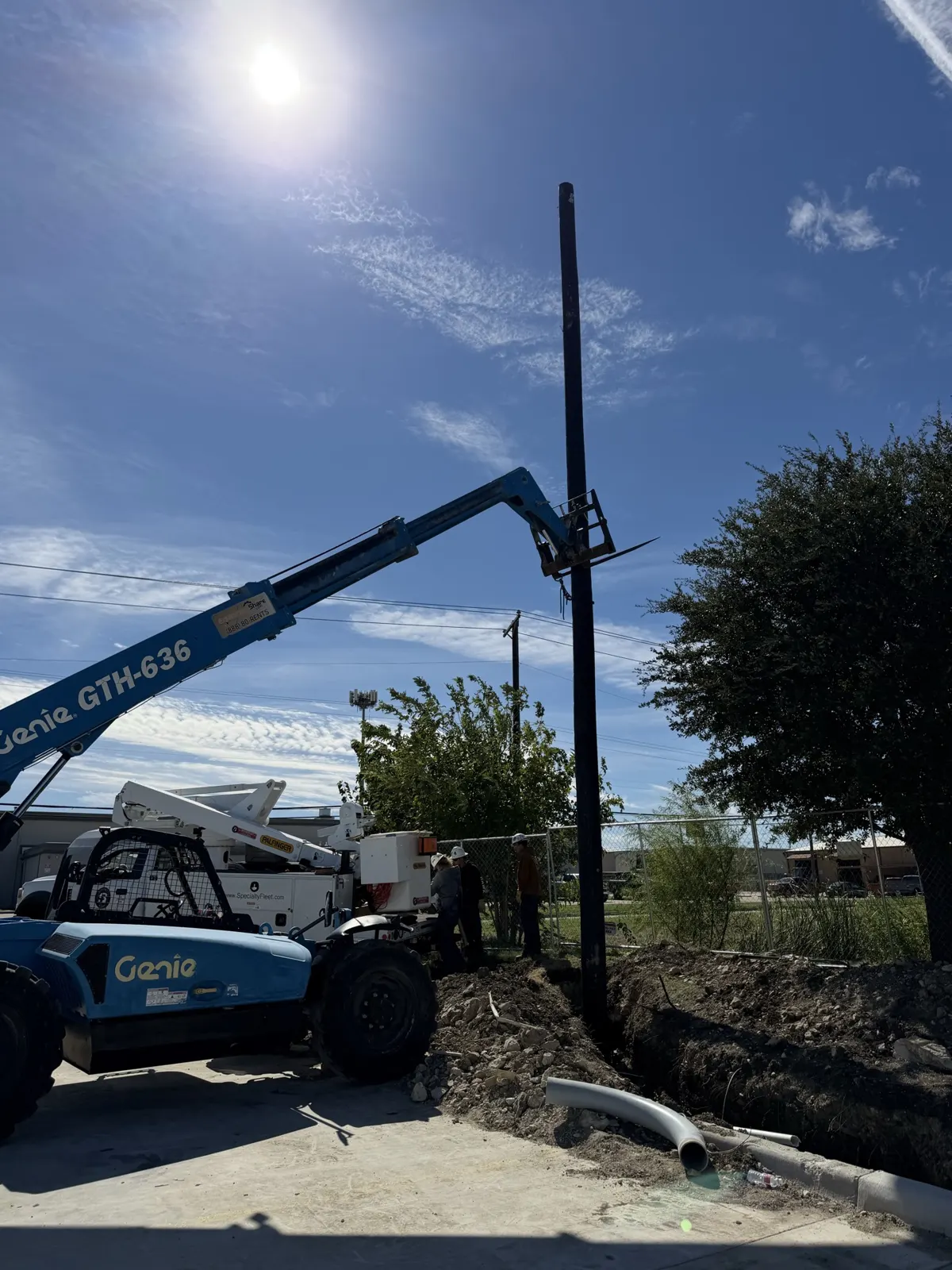 Telehandler lifting a utility pole beside a bucket truck at a Tesla charging station site in Saginaw, TX